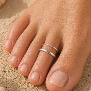 May include: A close-up of a foot on a sandy surface, adorned with two silver toe rings. The rings are simple bands, one above the other. The foot has a natural, neutral nail polish. A seashell is visible in the background.