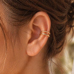 May include: Close-up of a gold ear cuff with two bands. The top band has a textured design, while the bottom band is smooth. The jewelry is worn on the upper ear cartilage.