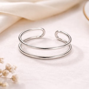 Può includere: Un braccialetto a polsino aperto color argento con un design a doppia fascia. Il braccialetto è realizzato in metallo lucido e liscio e poggia su una superficie bianca con un morbido tessuto color crema sullo sfondo. Un piccolo rametto di fiori secchi si trova nell'angolo in basso a sinistra.
