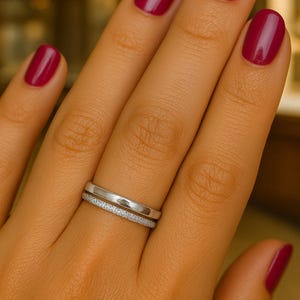 Könnte beinhalten: Zwei silberne Ringe, einer mit einem Band aus kleinen, funkelnden Steinen, werden auf einer Hand mit lackierten Nägeln gezeigt. Die Nägel sind in einem tiefen Rot lackiert, was einen Kontrast zu den Silberringen bildet.