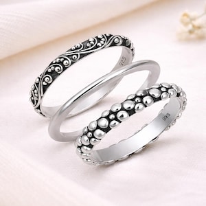 Peut inclure: Trois bagues en argent sur fond blanc. L'une présente un motif tourbillonnant orné, une autre est une simple bande, et la troisième a un motif texturé et pointillé. Les bagues sont probablement en argent sterling, comme l'indique le poinçon "925".