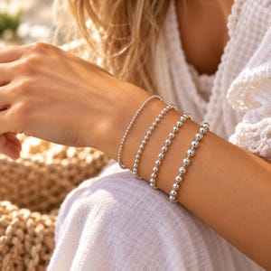 May include: A set of four silver-toned beaded bracelets worn on the wrist. The bracelets feature varying sizes of polished spherical beads, ranging from very small, fine beads to larger, prominent metallic spheres, creating a layered appearance.