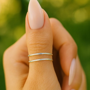 May include: A close-up of a hand wearing two thin, silver rings on the thumb. The rings are simple bands. The nails are long and painted with a light pink polish. The background is a blurred green.