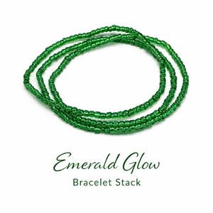May include: Three strands of emerald green beaded bracelets are displayed on a white background. The bracelets are made of small, round beads. The text "Emerald Glow Bracelet Stack" is below the bracelets.