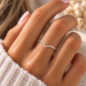 May include: A polished silver-toned ring featuring a simple V-shaped chevron design. The band is thin and smooth, showcasing a metallic finish that curves into a point at the center, resting on a finger with a clean and understated appearance.