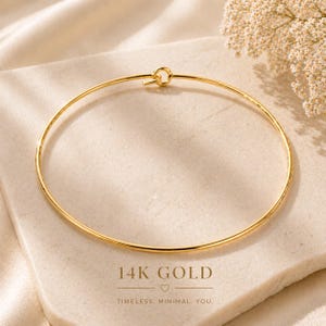 Può includere: Un bracciale rigido circolare sottile in oro 14 carati con un semplice meccanismo di chiusura a gancio. Il gioiello è lucidato a specchio e riflette la luce lungo la sua superficie liscia e arrotondata. Il testo sottostante recita 14K GOLD.