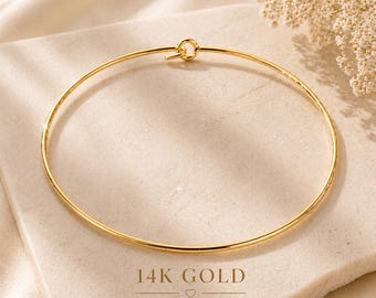 May include: A thin, circular 14K gold bangle bracelet featuring a small, integrated hook-and-eye closure mechanism. The text 14K GOLD is printed below the jewelry, followed by a small heart icon and the text TIMELESS. MINIMAL. YOU.