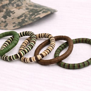 May include: Five stacked bracelets in shades of green, brown, and tan, with a camouflage-patterned cap in the background. The bracelets are made of small, flat, circular beads. The bracelets are approximately 2.5 inches in diameter.