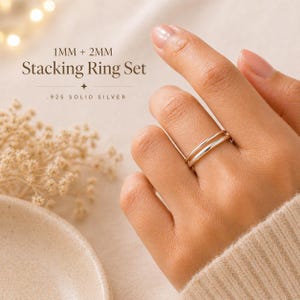May include: A close-up of a hand wearing a set of two silver stacking rings. The rings are 1mm and 2mm in width. The text in the image reads "1MM + 2MM Stacking Ring Set .925 SOLID SILVER".