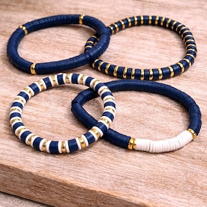 Puede incluir: Cuatro pulseras de cuentas en azul marino, blanco y dorado. Las pulseras están hechas de pequeñas cuentas redondas y planas. Algunas son de color azul marino liso, mientras que otras tienen detalles blancos y dorados. Se exhiben sobre una superficie de madera.