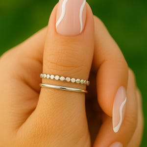 Puede incluir: Una mano con dos anillos de plata, uno con pequeños detalles circulares y el otro una banda lisa. Las uñas están pintadas con una base rosa claro y líneas onduladas blancas. El fondo es un verde borroso.