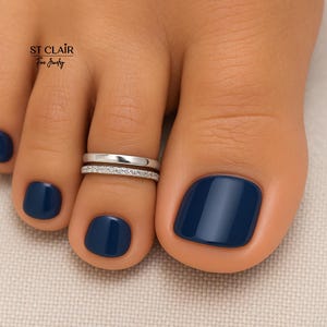 May include: Close-up of a foot with two silver toe rings. The top ring is plain, while the bottom ring is adorned with small, sparkling stones. The toenails are painted a deep navy blue. The image includes the text "ST CLAIR Fine Jewelry".
