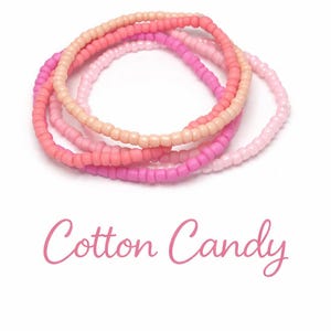 May include: Four beaded bracelets in shades of pink and peach, arranged on a white background. The bracelets are made of small, round beads. The text "Cotton Candy" is written in a cursive font below the bracelets.