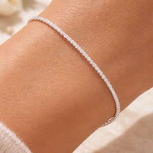 May include: A delicate silver bracelet, adorned with numerous small, sparkling stones, is shown on a person's wrist. The bracelet's design is simple yet elegant, reflecting light and adding a touch of glamour. The bracelet is a fine piece of jewelry.
