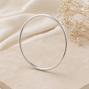 May include: A simple, silver-colored bangle bracelet. The circular bracelet is smooth and shiny, reflecting light. The jewelry is set against a neutral background with soft, natural lighting. The bangle is a classic piece of jewelry.