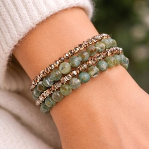 May include: A stack of four beaded bracelets. The bracelets feature a mix of green and gold beads. The green beads have a marbled appearance. The gold beads are small and faceted. The bracelets are worn on a wrist, with a white sweater in the background.
