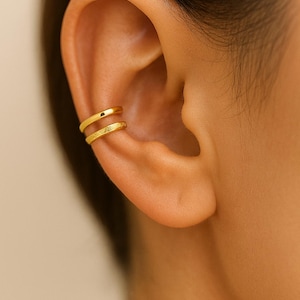 Könnte beinhalten: Nahaufnahme eines goldenen Ohrschmucks mit zwei Bändern. Das obere Band ist glatt, während das untere Band eine strukturierte, gehämmerten Oberfläche hat. Der Schmuck wird am Ohr getragen.