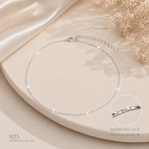 Puede incluir: Un delicado collar de plata de ley con un diseño de corte de diamante para un brillo extra. El collar tiene una extensión de 2,54 cm y está etiquetado como "925 STERLING SILVER". La joya se muestra sobre una superficie neutra con un fondo suave y texturizado.