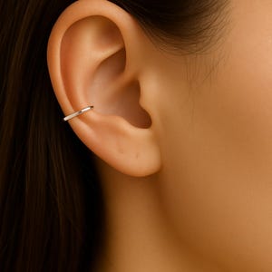 May include: A silver ear cuff is shown on the upper ear. The jewellery is a simple, thin band that curves around the ear's cartilage. The cuff is a minimalist design, reflecting a modern aesthetic. The ear cuff is a type of earring.