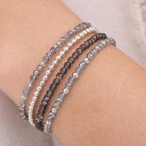 May include: A collection of beaded bracelets in shades of gray, silver, and black. The bracelets are made of small, round beads and are stacked on a wrist. The bracelets are a mix of crystal and metallic beads.