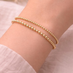 Pode incluir: Duas pulseiras de contas douradas. As pulseiras são feitas de pequenas contas redondas douradas. Uma pulseira tem contas maiores, enquanto a outra tem contas menores. As pulseiras são usadas no pulso.