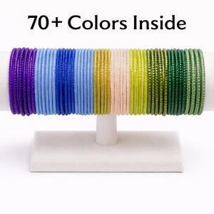 May include: A display of colorful bangles in shades of purple, blue, green, and gold, arranged on a white stand. The text "70+ Colors Inside" is displayed above the bangles.