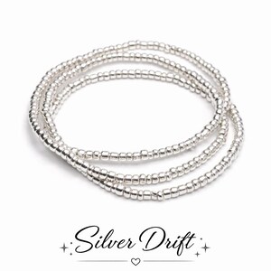 Pode incluir: Três pulseiras de contas prateadas, feitas de pequenas contas redondas e brilhantes, sobre um fundo branco. As pulseiras estão dispostas em forma circular. O texto "Silver Drift" está escrito em itálico abaixo.