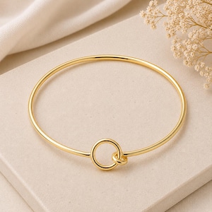 May include: A gold-colored bangle bracelet with a simple, elegant design. The bracelet features a circular band with a smaller circle attached at the front. The jewelry is displayed on a neutral surface with a soft, blurred background.