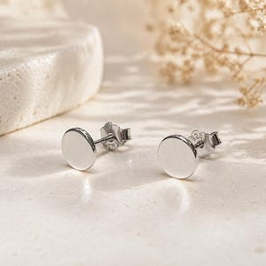 May include: A pair of silver stud earrings with a simple, round design. The earrings are made of polished metal and are displayed on a light-colored surface. The earrings are a minimalist style, suitable for everyday wear.