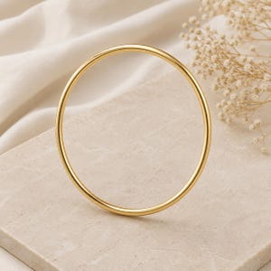 May include: A gold-colored bangle bracelet. The circular bracelet is displayed on a neutral-toned surface, with a sprig of dried flowers in the background. The bangle has a smooth, polished finish and a simple, elegant design.