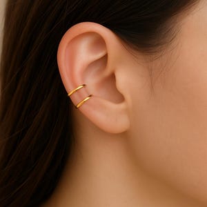 May include: A gold-colored ear cuff featuring a double-band design. The jewelry is worn on the ear cartilage, creating a minimalist and modern look. The cuff is made of a shiny, metallic material.