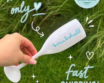 Può includere: Un flûte da champagne bianco con la parola bombshell scritta in corsivo color ottanio. Il bicchiere presenta scritte che indicano hand wash only, small business e fast shipping, insieme a icone decorative di cuori e stelle.