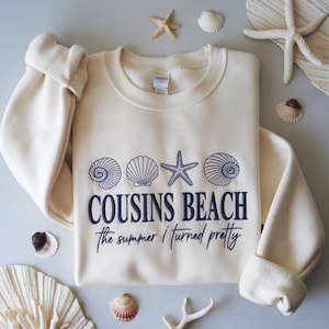 May include: Cream-colored sweatshirt with the text "COUSINS BEACH" and "the summer I turned pretty" in navy blue. The design includes seashell and starfish graphics. The sweatshirt is surrounded by seashells and starfish.