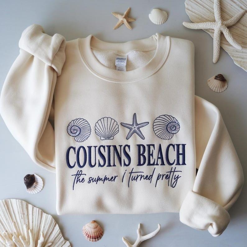 May include: Cream-colored sweatshirt with the text "COUSINS BEACH" and "the summer I turned pretty" in navy blue. The design includes seashell and starfish graphics. The sweatshirt is surrounded by seashells and starfish.