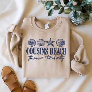 May include: A beige sweatshirt with the text "COUSINS BEACH" and "the summer I turned pretty" in blue. The design includes blue seashell and starfish graphics. Brown sandals are visible in the lower left corner.