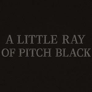 May include: Dark fabric with the embroidered words "A LITTLE RAY OF PITCH BLACK" in grey. The text is centered and in a serif font, creating a simple, elegant design.