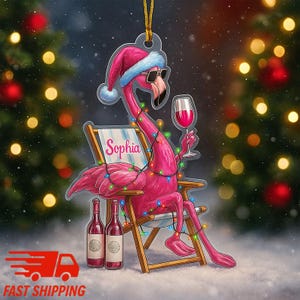 May include: A pink flamingo Christmas ornament wearing a Santa hat and sunglasses, holding a glass of wine, and sitting in a beach chair. The chair has the name "Sophia" on it. The flamingo is decorated with fairy lights, and two wine bottles are at its feet.
