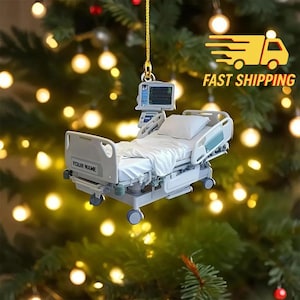 May include: A miniature hospital bed ornament with a monitor and the text "YOUR NAME". The bed is white and gray, with a white pillow and bedding. The ornament is hanging in front of a Christmas tree.