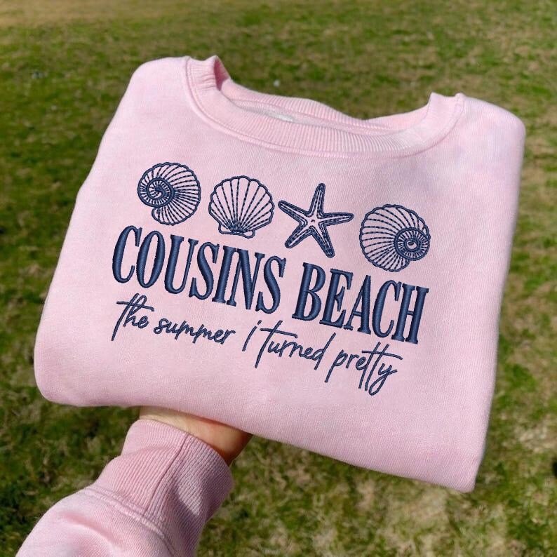 May include: A light pink sweatshirt with the text "COUSINS BEACH" and "the summer I turned pretty" in navy blue. The design includes seashell and starfish illustrations. A casual, beach-themed apparel item.