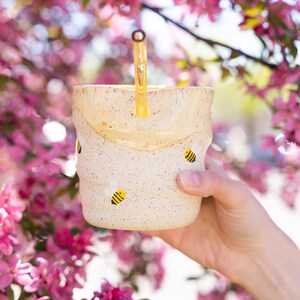 Peut inclure: Une tasse en céramique tachetée avec un glaçage jaune semblable à du miel et des illustrations d'abeilles. Une paille jaune dépasse de la tasse. Le fond présente des fleurs roses.
