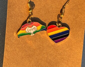 Może przedstawiać: Złote kolczyki z tęczowym sercem i tęczową dłonią z białym napisem "love". Kolczyki są prezentowane na brązowej powierzchni.