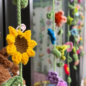 May include: A hanging decoration featuring multiple vertical strands of crocheted flowers in various colors including yellow, orange, blue, purple, and pink, with green crocheted stems and leaves, and a small pink plastic stitch marker attached.