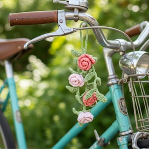 Op de afbeelding: Een gehaakte bloemenfietsversiering met drie rozen in roze en koraal, met groene bladeren en stelen. De versiering hangt aan het stuur van een vintage turquoise fiets met bruine lederen details.