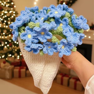 May include: A handmade crochet bouquet of blue flowers with yellow centers and green leaves. The flowers are arranged in a white, cone-shaped holder. The background includes a Christmas tree and wrapped gifts.