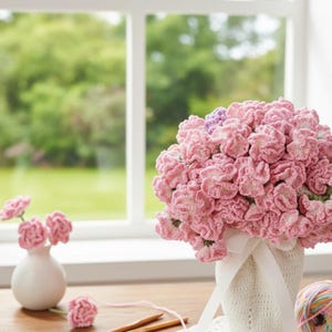 May include: A large bouquet of pink crocheted flowers in a white vase with a white ribbon. A small white vase with three pink crocheted flowers is on the left. Wooden knitting needles and yarn are on the wooden table.