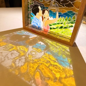 Puede incluir: Un marco de madera cuadrado que contiene un panel de vidriera que representa a una pareja formando un corazón con las manos frente a un paisaje de colinas y árboles. La obra de vidrio proyecta una imagen colorida y translúcida debajo.