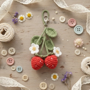 Könnte beinhalten: Ein handgefertigter Kirsch-Schlüsselanhänger aus Häkelarbeit mit roten Kirschen, weißen Blütenakzenten und grünen Blättern. Der Schlüsselanhänger liegt auf einem Holzgrund mit Knöpfen, Spitze und Blumen.