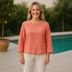 Puede incluir: Una blusa de manga larga de color coral con cuello redondo y mangas tres cuartos. La blusa se combina con pantalones beige claro. El fondo presenta una piscina y vegetación.