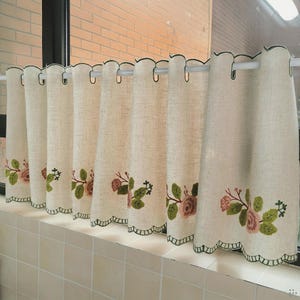 Könnte beinhalten: Beige Küchenvorhänge aus Leinen mit gewelltem Rand und gesticktem Blumendesign. Die Vorhänge haben eine dunkelgrüne Bordüre und sind an einer weißen Stange aufgehängt. Die Blumenstickerei weist rosa und grüne Farben auf.