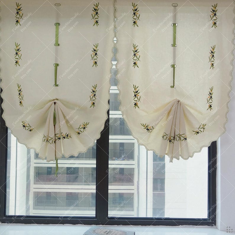 May include: Cream-colored window curtains with olive branch embroidery. The curtains have a scalloped edge and are gathered at the bottom. Green ribbon accents are visible. The curtains are hanging in front of a window.
